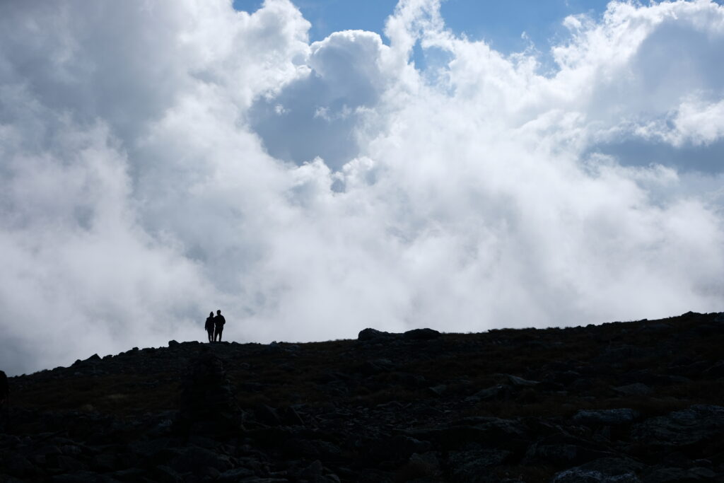 Two people standing, silhouetted on a ridge with sun and blue sky breaking through clouds